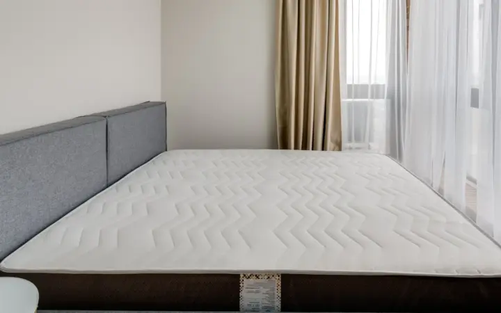 A bare quilted mattress on a bed frame, lit by daylight from a window on the right